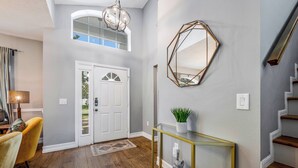 Un hall d’entrée moderne avec un plancher en bois, une porte blanche, une console en verre ornée d’une plante et un miroir géométrique accroché au mur.