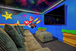 A room with a sofa, ottoman, and a wall mural featuring Mario.
