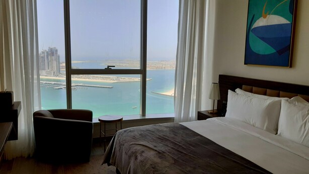 A hotel room with a large window offering a view of a coastal cityscape, a bed, a chair, and a bedside table. - Dubai Dream - Hotel Suite - Breathtaking view of Palm Jumeira +++ (Dubai)