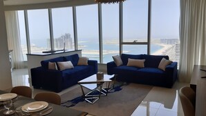A modern living room with two blue sofas, a small coffee table, and a dining area with a table set for four. Large windows offer a view of a cityscape and a body of water. - Dubai Dream - Hotel Suite - Breathtaking view of Palm Jumeira +++ (Dubai)