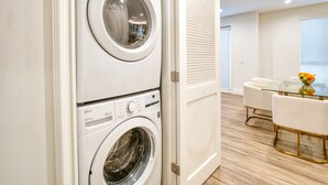 A laundry room with two stacked washers and dryers, a glass table with a fruit bowl, and a white door with blinds.