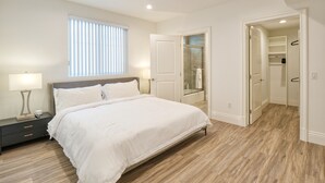 A bedroom with a large bed, bedside lamps, a window with blinds, a walk-in closet, and a shower area.