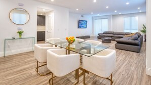 A modern living room with a glass-top dining table, white chairs, a gray sofa, and a flat-screen TV mounted on the wall.