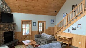 A cozy living room with a stone fireplace, a large gray sofa, a wooden coffee table, and a wooden staircase. - 4bdrm Chalet-close to downtown & outdoor adventure (Bethel)