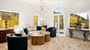 A spacious living room with a large chandelier, a sofa set, a coffee table, and a view of autumn foliage through large windows. - Exquisite pet friendly home, 5 blks from Gondola with stunning views!! (Aspen)