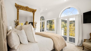 A bedroom with a large bed, a gold-framed mirror, and a balcony with a view of trees. - Exquisite pet friendly home, 5 blks from Gondola with stunning views!! (Aspen)