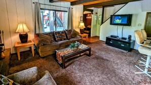 A cozy living room with a wooden ceiling, a large window, a television, and a sofa with patterned cushions.