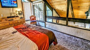 A bedroom with a sloped ceiling, a bed with a red and white bedspread, a television on the wall, and a window with blinds.