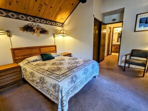 Ein Schlafzimmer mit einem hölzernen Kopfteil, einer blumigen Tagesdecke, zwei Nachttischlampen, einem Sessel und einer Tür zu einem anderen Raum.