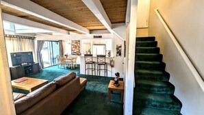 A cozy living room with a green carpet, a sofa, a coffee table, and a TV. There is a staircase to the right.