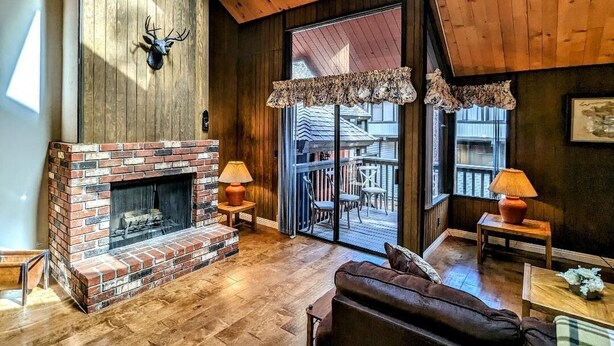 A cozy living room with a brick fireplace, wooden ceiling beams, and a large window overlooking a deck.