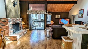 A cozy living room with a brick fireplace, wooden ceiling, and a large window overlooking a balcony.