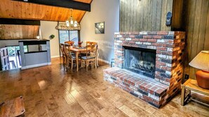 A rustic interior with a brick fireplace, wooden ceiling beams, a dining area with chairs, and a mounted deer head.