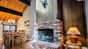 A cozy room with a brick fireplace, wooden ceiling beams, and a dining area with chairs and a table.