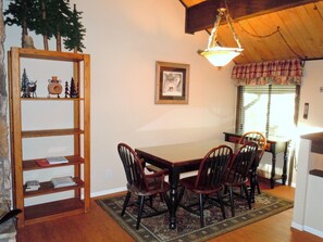 A dining area with a wooden table, four chairs, a chandelier, a bookshelf, and a window with curtains.