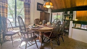 Un espace repas avec une table et des chaises en bois, une lampe suspendue et une vue sur l’extérieur grâce à de grandes fenêtres.
