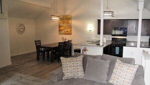 A modern living room with a gray sofa, a dining table with chairs, and a kitchen area with dark cabinets and a microwave.