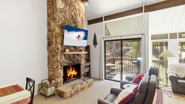 A living room with a stone fireplace, a television mounted above it, a cozy sofa, and a view through a sliding glass door to an outdoor deck.