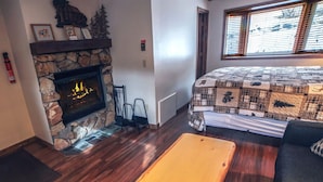 Una habitación acogedora con chimenea de piedra, una cama con colcha, suelo de madera y una ventana con persianas.