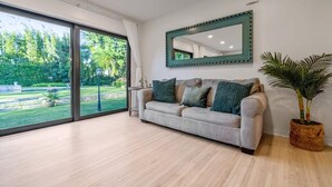 A modern living room with a large sliding glass door leading to a garden, a sofa with cushions, a mirror, and a potted plant. - Garden House, Pool in Lush Peaceful Homestead (Homestead)