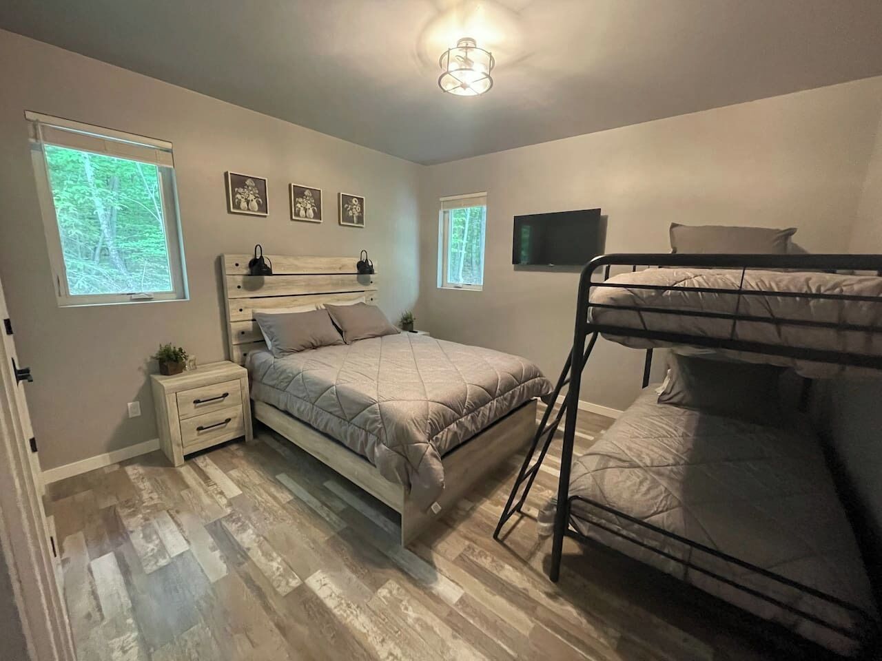 Une chambre avec un lit superposé, un lit simple, une table de chevet, une télévision et une fenêtre donnant sur des arbres.