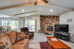 A living room with a stone fireplace, a television, a couch, and a dining area with chairs and a table. - Furnished Deck & Fire Pit: Sherwood Forest Cabin (Twain Harte)