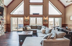 A spacious living room with a large couch, a coffee table, and a dining area. There are large windows allowing natural light, and a sign on the wall that reads "DO NOT FEED THE BEARS".