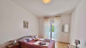 A bedroom with a single bed, a bedside table, a lamp, a window with curtains, and a view of greenery outside. - Holiday apartment with terrace, air conditioning and garden (Dramalj)