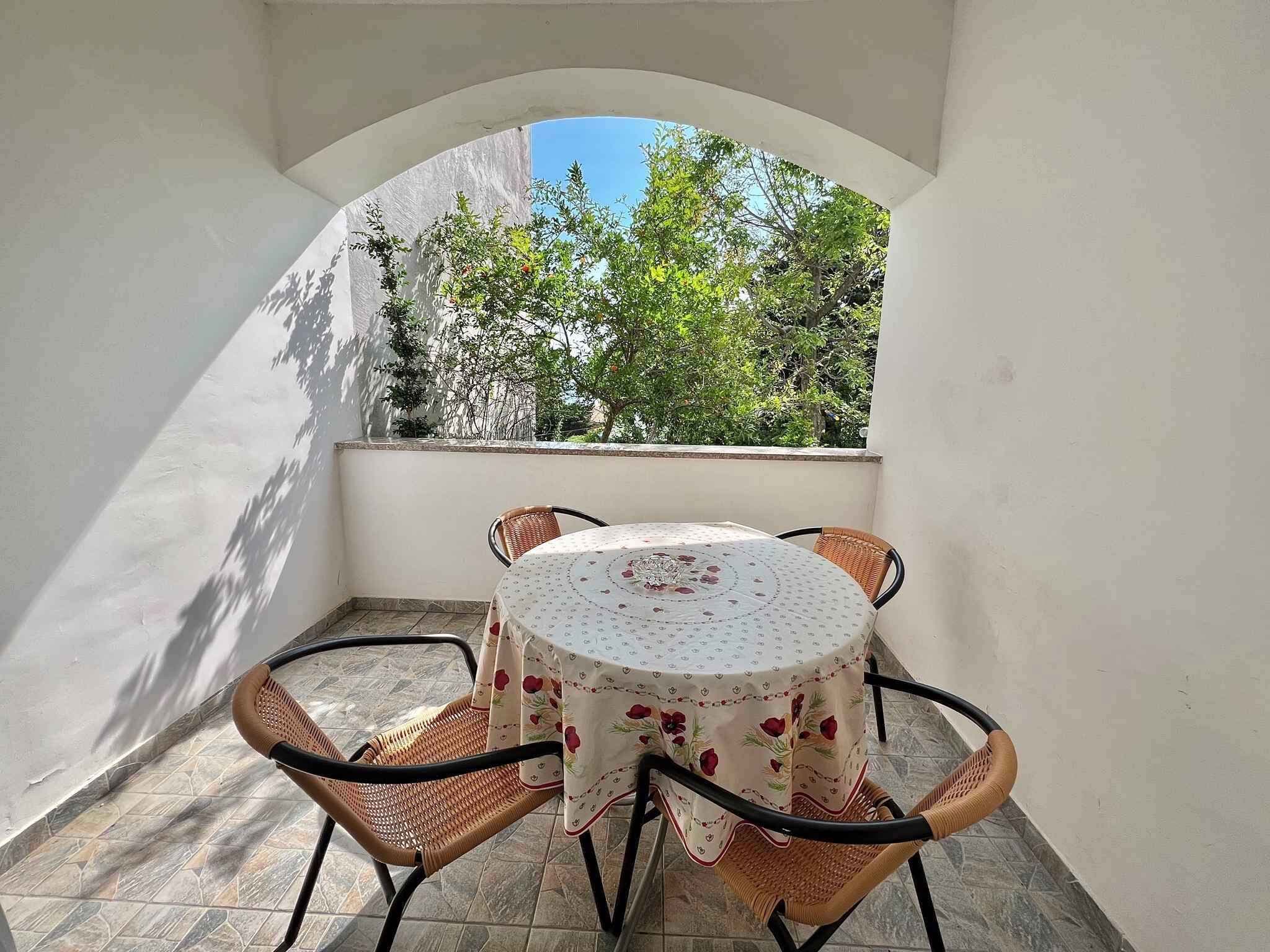 Un pequeño patio al aire libre con una mesa redonda y cuatro sillas de mimbre, rodeado por un muro blanco y una ventana arqueada con vistas a un jardín.