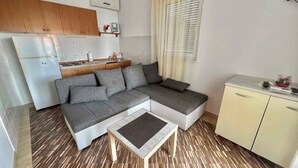 A compact living space with a grey sectional sofa, a small wooden coffee table, a kitchenette with a fridge and stove, and a window with blinds. - Holiday apartment with terrace and air conditioning (Dramalj)