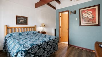 A bedroom with a bed, a nightstand, a lamp, a framed picture of flowers, a clock, and a door.