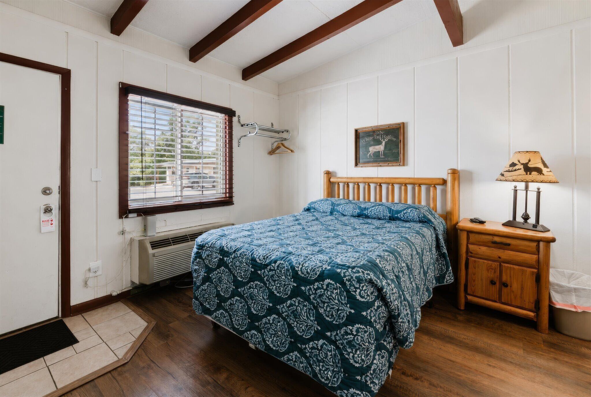 Un dormitorio con cama, mesita de noche, lámpara, ventana con persianas, puerta y un cuadro colgado en la pared.