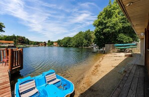 A lakeside dock with a boat and chairs, a sandy beach, and a wooden deck.