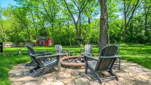 Een vuurplaats van natuursteen omringd door Adirondack-stoelen in een weelderige achtertuin, met een rode schuur op de achtergrond.