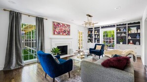 Een moderne woonkamer met een open haard, blauwe fauteuils, een grijze bank en een boekenkast vol decoratieve objecten.