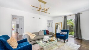 Een moderne woonkamer met een glazen salontafel, blauwe fauteuils, een grijze bank en een gouden kroonluchter.