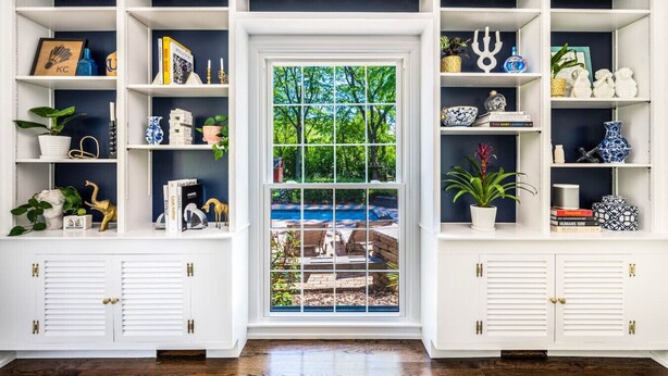 Een kamer met witte inbouwplanken, een groot raam met uitzicht op een zwembad en groen, en een houten vloer.