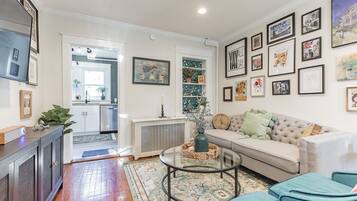 A living room with a sofa, a coffee table, and a TV. There are framed pictures on the wall and a plant on the floor.