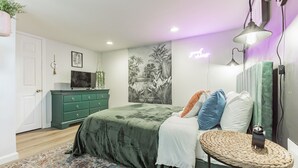 Un dormitorio con cama, cómoda, espejo, lámpara de pared y taburete tejido.