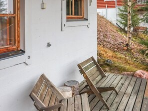 A wooden deck with two chairs and a small table, a white building with windows, and a natural landscape with trees and a hill in the background.