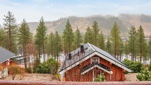 Una cabaña con techo inclinado, rodeada por un bosque y colinas al fondo.