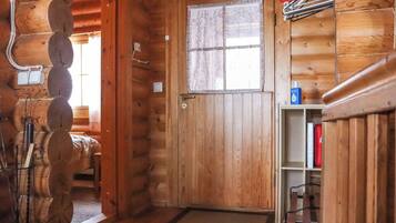 A wooden cabin interior with a door, a window with curtains, a shelf with items, and a hanging coat rack.