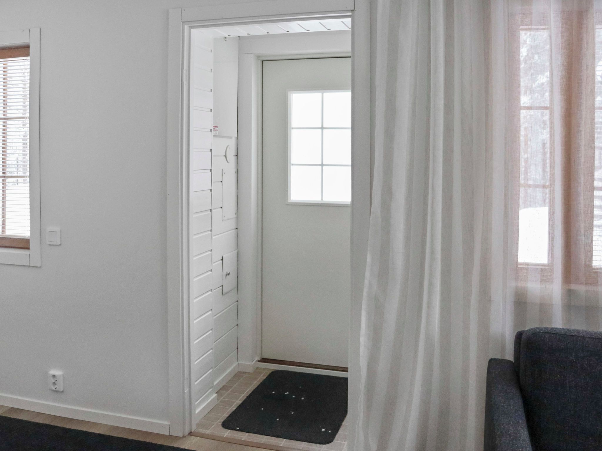 Ein Zimmer mit einer weißen Tür, einem Fenster mit Jalousien, einer schwarzen Fußmatte und einem grauen Sofa.