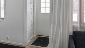 Een kamer met een witte deur, een raam met zonwering, een zwarte deurmat en een grijze bank.