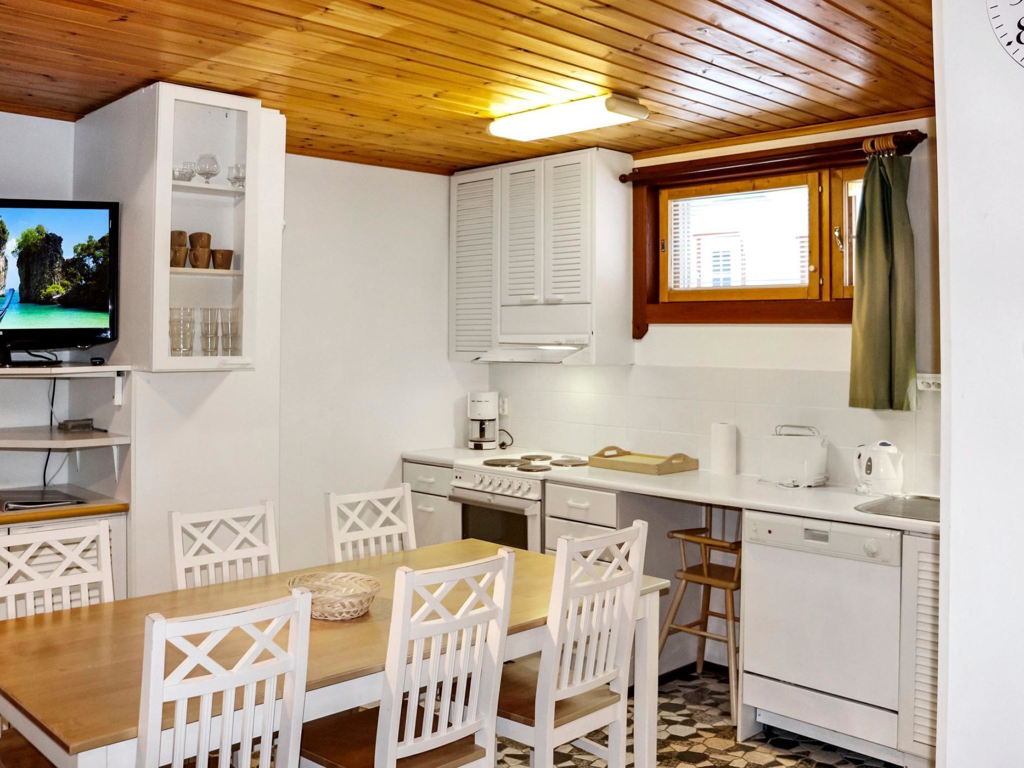 Une cuisine de taille réduite avec des meubles blancs, une table à manger en bois et un téléviseur posé sur une étagère.