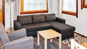 A modern living room with a grey sectional sofa, a small wooden table, and a grey armchair. There are two windows with blinds, a floor lamp, and a wooden door. - Alppikylä 8b by Interhome (Hyrynsalmi)