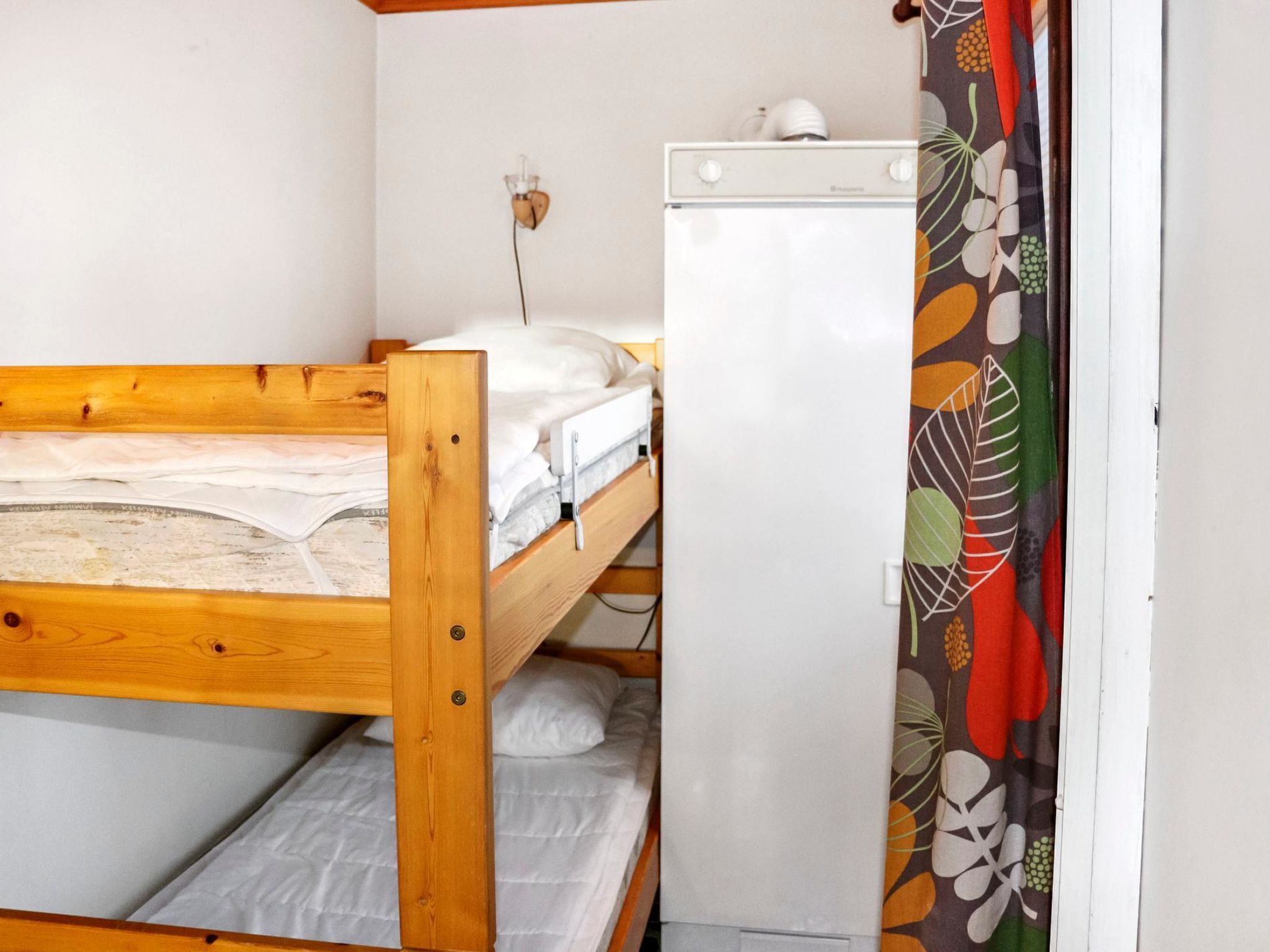 Una habitación pequeña con una cama individual de madera, una pared blanca y un refrigerador blanco.