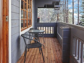 Una terraza de madera con dos sillas y una mesita, una ventana con marcos de madera y vistas a árboles.