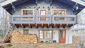 Une cabane à deux étages dotée d’un balcon, de grandes fenêtres et d’une pile de bois de chauffage devant l’entrée.