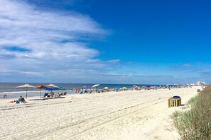 En strand med folk som kopplar av under parasoller, en sandstrand och en klar himmel.